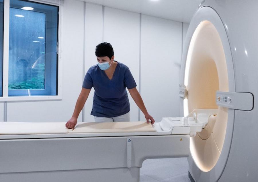 Magnetresonanztomografie-Gerät, Seitenansicht, Mitarbeiterin mit Mund-Nasen-Schutz am Gerät stehend