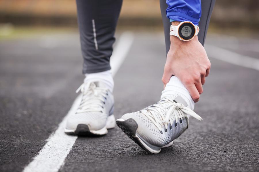 Füße eines Läufers in Sportschuhen auf Laufstrecke, linker Fuß umgeknickt, linke Hand am Sprunggelenk