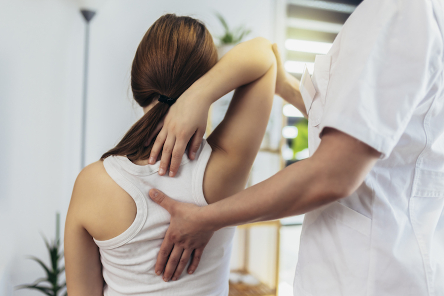 Frau, von hinten zu sehen, macht Rückenübung unter Anleitung einer Physiotherapeutin