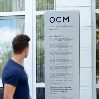 Junger Patient, seitlich zu sehen, blickt auf das Eingangsschild der OCM Orthopädische Chirurgie München