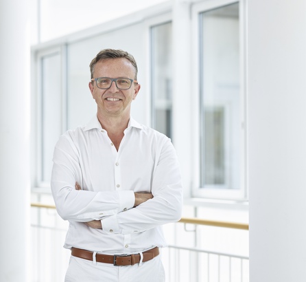Halbportrait Prof. Dr. Robert Hube von der OCM München, er lächelt, trägt weiße Kleidung, befindet sich im Treppenhaus, weißer Hintergrund